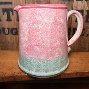 May include: A large, handmade ceramic pitcher with a pink and green speckled design. The pitcher has a rounded base, a handle, and a spout. The top and handle are trimmed in a darker pink. The pitcher sits on a wooden surface.