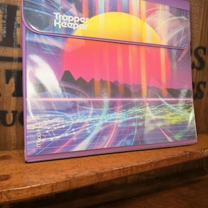 May include: Purple Trapper Keeper binder with a retro sunset design. The binder features a vibrant graphic of a yellow sun, pink and purple sky, and a grid pattern. The text "Trapper Keeper" is visible on the front.