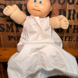 May include: A vintage Cabbage Patch Kid doll with a round, pale face, blue eyes, and a tuft of blonde hair. The doll is wearing a white dress with lace trim. The doll is sitting on a wooden bench.