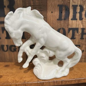 Puede incluir: Figura de caballo de cerámica blanca en pose de encabritado. El caballo está esculpido con detalles y se apoya en una base decorativa. La figura es de color blanco brillante y se exhibe sobre una superficie de madera.