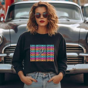 May include: Black crew neck sweatshirt with the text "SAN ANTONIO SPURS" in a repeating, colourful design. The San Antonio Spurs logo is visible within the text. The sweatshirt is worn with blue jeans.