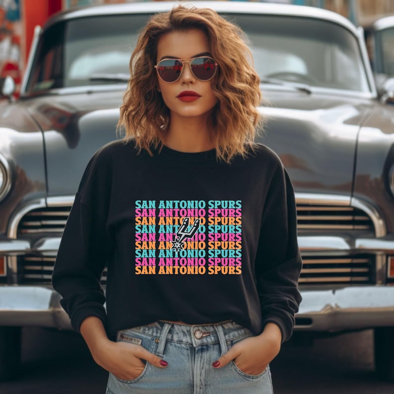 May include: Black crew neck sweatshirt with the text "SAN ANTONIO SPURS" in a repeating, colourful design. The San Antonio Spurs logo is visible within the text. The sweatshirt is worn with blue jeans.