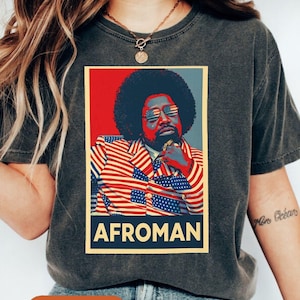 May include: Dark gray t-shirt featuring a graphic of a man with an afro, wearing sunglasses with an American flag design, and a jacket with an American flag design. The word "AFROMAN" is printed in white letters on a blue banner.