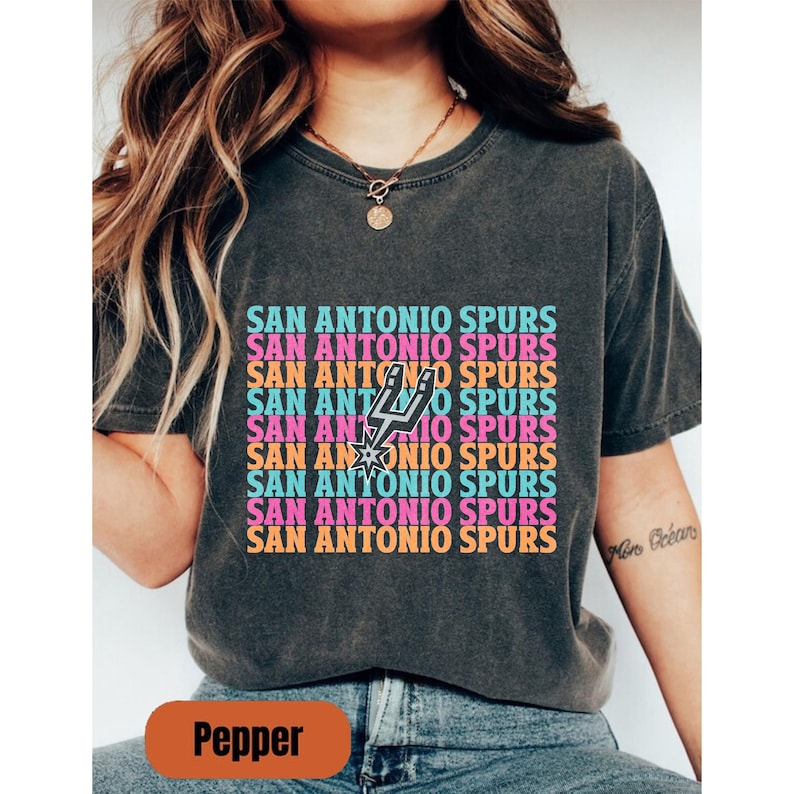 May include: Dark grey t-shirt with the words "SAN ANTONIO SPURS" repeated in a colourful pattern, including the team logo. The text is in pink, orange, and blue. The shirt has a relaxed fit.