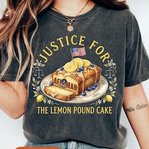 Puede incluir: Camiseta gris oscuro con el texto "JUSTICE FOR THE LEMON POUND CAKE". El gráfico presenta un bizcocho decorado con limones, arándanos y una pequeña bandera estadounidense. La palabra "Pepper" está impresa en un óvalo naranja.