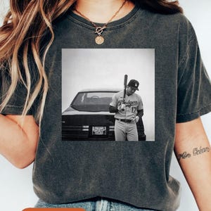 May include: Dark gray t-shirt with a black and white image of a baseball player holding a bat. The player is wearing a Dodgers uniform and standing near a black car. The word "Pepper" is printed on the bottom.