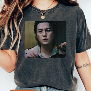 May include: Dark gray t-shirt with a square image of a person wearing glasses. The image is centered on the shirt. The word "Pepper" is printed on an orange oval at the bottom of the shirt.