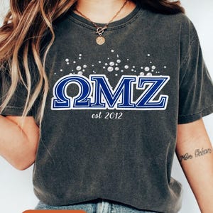 Pode incluir: Uma camiseta cinza escuro com as letras "ΩMZ" em azul e branco, com "est 2012" abaixo. A camiseta tem um visual desgastado e decote redondo. A palavra "Pepper" está em uma etiqueta marrom.