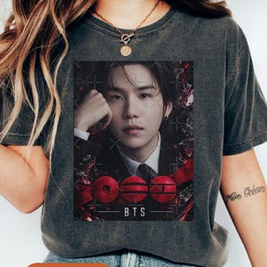 May include: Dark gray t-shirt featuring a portrait of a person with long dark hair. The graphic includes the text "BTS" and three red circular designs. The shirt is made of a soft material.