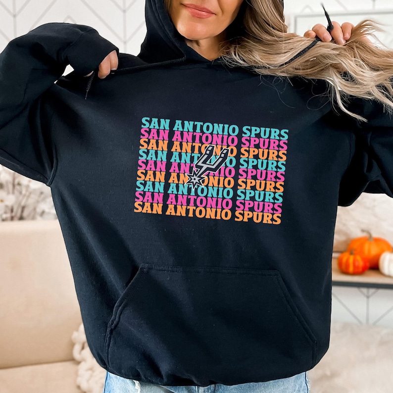 May include: Black hoodie with the text "SAN ANTONIO SPURS" in a repeating, colourful design. The design includes the Spurs logo. The hoodie has a drawstring hood and a front pocket.