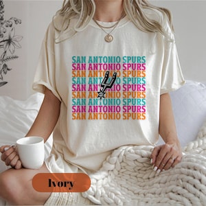 May include: Cream-coloured t-shirt with the text "SAN ANTONIO SPURS" in a repeating pattern of teal, pink, orange, and purple. The shirt features the Spurs logo in black and white. The model is holding a white mug.
