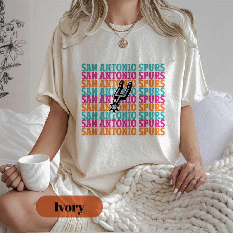 May include: Cream-coloured t-shirt with the text "SAN ANTONIO SPURS" in a repeating pattern of teal, pink, orange, and purple. The shirt features the Spurs logo in black and white. The model is holding a white mug.