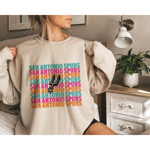 May include: Beige sweatshirt featuring the text "SAN ANTONIO SPURS" in a repeating, colourful pattern. The Spurs logo is in the centre. The sweatshirt has long sleeves.