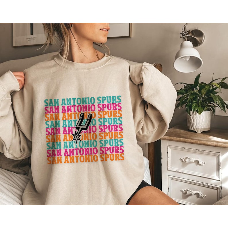 May include: Beige sweatshirt featuring the text "SAN ANTONIO SPURS" in a repeating, colourful pattern. The Spurs logo is in the centre. The sweatshirt has long sleeves.
