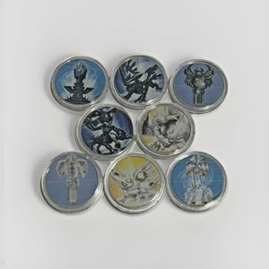 May include: A collection of eight circular game tokens with silver rims. Each token features a different colorful fantasy character or object design set against a blue or yellow background. The tokens are arranged on a white surface.