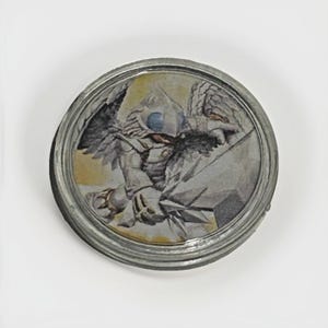May include: A round, metal-rimmed object featuring a detailed illustration of a winged creature with a blue orb. The artwork is set against a yellow and gray background, with the creature's form rendered in shades of gray and white.