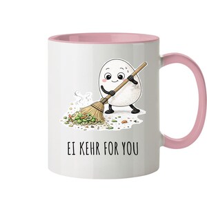 Könnte beinhalten: Weiße Keramik-Tasse mit rosa Innenraum und Henkel. Die Tasse zeigt einen Cartoon-Ei-Charakter, der Essen mit einem Besen zusammenkehrt. Der Text "EI KEHR FOR YOU" ist darunter gedruckt.