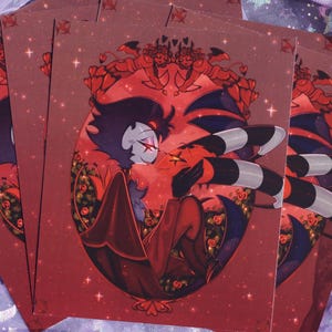 May include: Several art prints featuring a red and black cartoon character with a striped tail and horns. The character is set against a red background with floral and star details. The prints are stacked on a purple fabric.