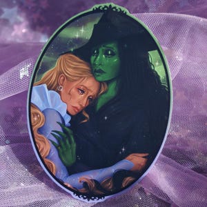 May include: An oval-shaped artwork depicts two figures embracing. One figure has green skin and wears a black hat, while the other has blonde hair and a blue dress. The artwork is set against a background of green and purple hues.