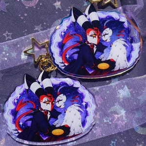 May include: Two acrylic keychains featuring a colorful illustration of two animated characters in a dark, starry, purple-toned setting. Each keychain has a gold-colored star-shaped clasp and depicts a red-skinned character and a white-furred character.