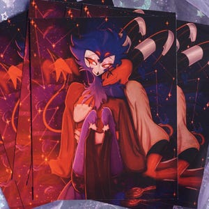 May include: A stack of art prints featuring a stylized illustration of a character with blue and red tones. The artwork has a celestial background with constellations and stars. The character is wearing a red cloak.