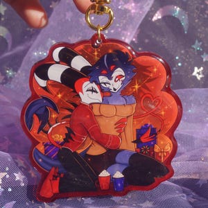 May include: A red and orange acrylic charm featuring two cartoon characters embracing. One character has black and white striped ears, and the other is blue. The charm has a gold-colored keyring and is decorated with hearts and gift boxes.