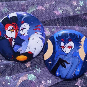 May include: Two circular pin-back buttons featuring colorful character illustrations. The left button depicts two figures in a close embrace with red, white, and blue tones. The right button shows a single blue-toned character with red eyes.