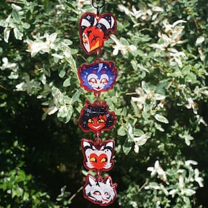 May include: A vertical hanging ornament featuring five connected acrylic charms, each displaying a colourful cartoon character head. The characters include a red and white imp, a blue owl-like figure, a red-skinned character, a white and red imp, and a white wolf.