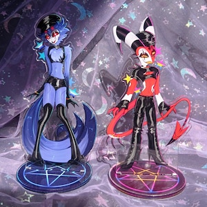 May include: Two acrylic character standees featuring stylized figures. The left figure is blue with a black hat and outfit. The right figure is red, black, and white with horns and a tail. Both stand on circular bases with a glowing pentagram design.