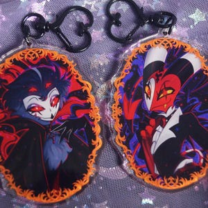 May include: Two acrylic keychains featuring cartoon characters with red eyes and black outfits. The keychains have an orange decorative border and black clasps. One character has blue fur and the other has red and white skin.