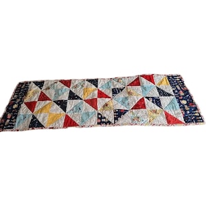 May include: A colorful quilted table runner featuring a patchwork design of triangles in red, yellow, blue, and white. The runner has a navy blue border with a whimsical pattern.
