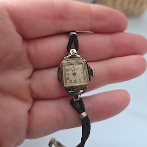 May include: Vintage silver-toned wristwatch with a square face and black cord band. The watch face has a cream-colored background with black numerals and hands. The watch has a small gold-toned crown and the word "WALER" is visible on the watch face.