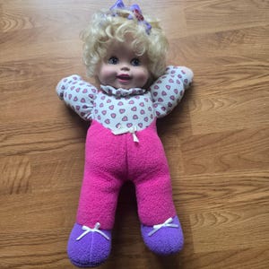 May include: A doll with curly blonde hair and a purple bow, featuring a white top with a pink heart pattern, a pink fleece-textured jumpsuit, and purple fleece-textured feet. The doll has blue eyes, a small white bow at the waist, and white ribbons.