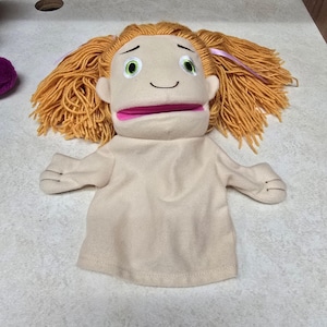 May include: A tan hand puppet with a smiling face, green eyes, and orange yarn hair. The puppet has a pink mouth and is wearing a tan shirt. The puppet's arms are extended.
