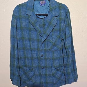 May include: A blue and green plaid blazer with two front pockets and three buttons. The blazer is hanging on a black hanger. The interior lining is a solid teal color.
