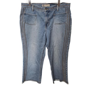 May include: Light blue denim jeans with decorative stitching down the sides and around the pockets. The jeans have a straight leg cut with a raw hem. The jeans are hanging on a metal hanger.