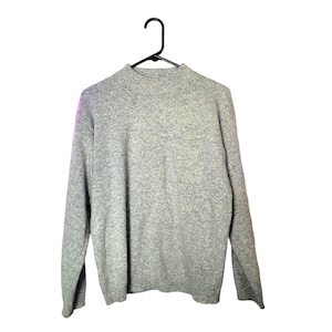 May include: A light gray sweater with a mock neck and long sleeves hangs on a black hanger. The sweater has a pocket on the front and is made of a soft, textured material. The background is white.