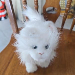 May include: A fluffy white cat figurine with blue eyes and a pink nose sits on a wooden chair. The cat has long, soft fur and is posed in a walking position. The chair has a light brown finish.