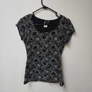 May include: Black short-sleeved top with a scoop neckline and a silver floral lace overlay. The top is hanging on a black hanger against a white background. The lace design covers the entire front and back of the top.