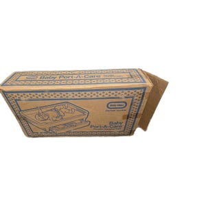 May include: A cardboard box for a Baby Port-A-Care portable changing table. The box is brown with blue and white accents. The box has an illustration of the changing table and the words "Baby Port-A-Care" printed on it.