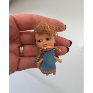 May include: A small doll with messy ginger hair, blue eyes, and long black eyelashes. The doll wears a light blue sleeveless dress featuring a gold glittery trim around the neckline and hem. The doll has a pale complexion and small arms.