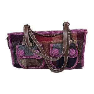 May include: A patchwork handbag with a mix of colors including purple, gray, burgundy, and brown. The bag has two front pockets with round, ribbed purple accents. It features brown leather straps and a chain detail, with a small lock closure.