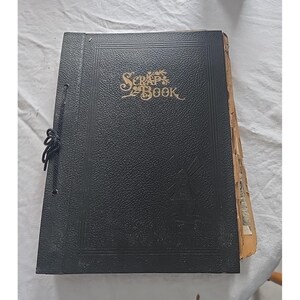 May include: A vintage black scrapbook with the word "SCRAP BOOK" in gold lettering on the cover. The cover has a textured appearance and is bound with a black cord. The pages are visible on the right side.