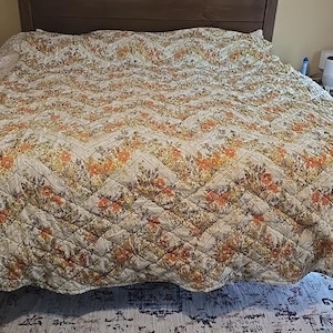May include: A bed covered with a floral patterned quilt. The quilt features a cream background with orange, yellow, and green floral designs. The bed is made up with a dark brown headboard and a patterned rug is visible beneath the bed.