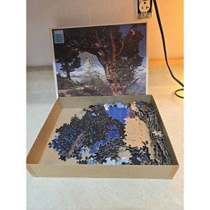 May include: A 1000-piece jigsaw puzzle with a landscape scene featuring a large tree and a snow-capped mountain. The puzzle pieces are in a cardboard box, partially assembled. The box has the text "1000 Pieces".