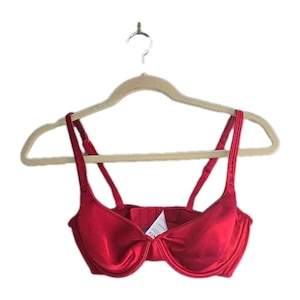 May include: A red satin bra with adjustable straps hangs on a beige hanger. The bra has underwire support and a hook-and-eye closure in the back. The bra is a solid, vibrant red color.