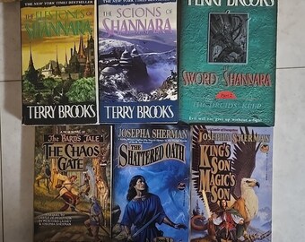 May include: A collection of fantasy novels by Terry Brooks and Josepha Sherman. The books feature colorful covers with titles like "The Elfstones of Shannara," "The Scions of Shannara," and "The Sword of Shannara." The books are displayed on a light brown surface.