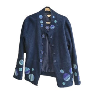 May include: A navy blue jacket with a unique design. The jacket features large, circular appliqués in shades of blue and green on the sleeves and around the collar. It has a button-up front and a tailored fit, perfect for layering.