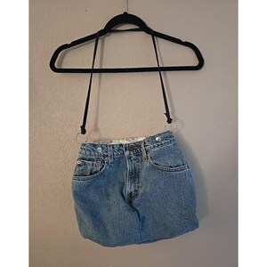 May include: A repurposed denim skirt handbag, featuring a light blue wash. The bag is suspended from a black hanger with black straps and silver metal rings. The skirt's pockets and button closure are visible.