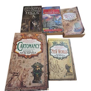May include: A collection of fantasy novels with various covers. Titles include "Beneath Vaulted Hills," "The Shadow Roads," "Secret Atlas," "Cartomancy," and "The New World." The books feature illustrations and text.
